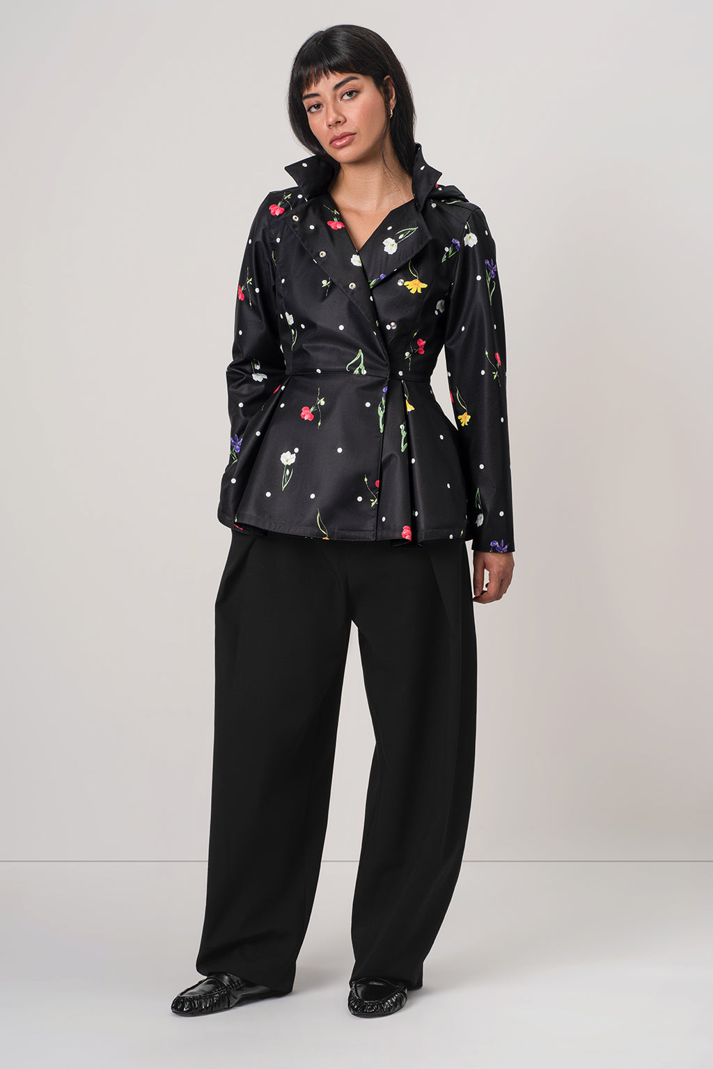 A model wearing RainSisters black long-sleeve double breasted jacket Cosmic Meadow with a colorful floral print and black wide-leg trousers, standing against a light gray studio background.
