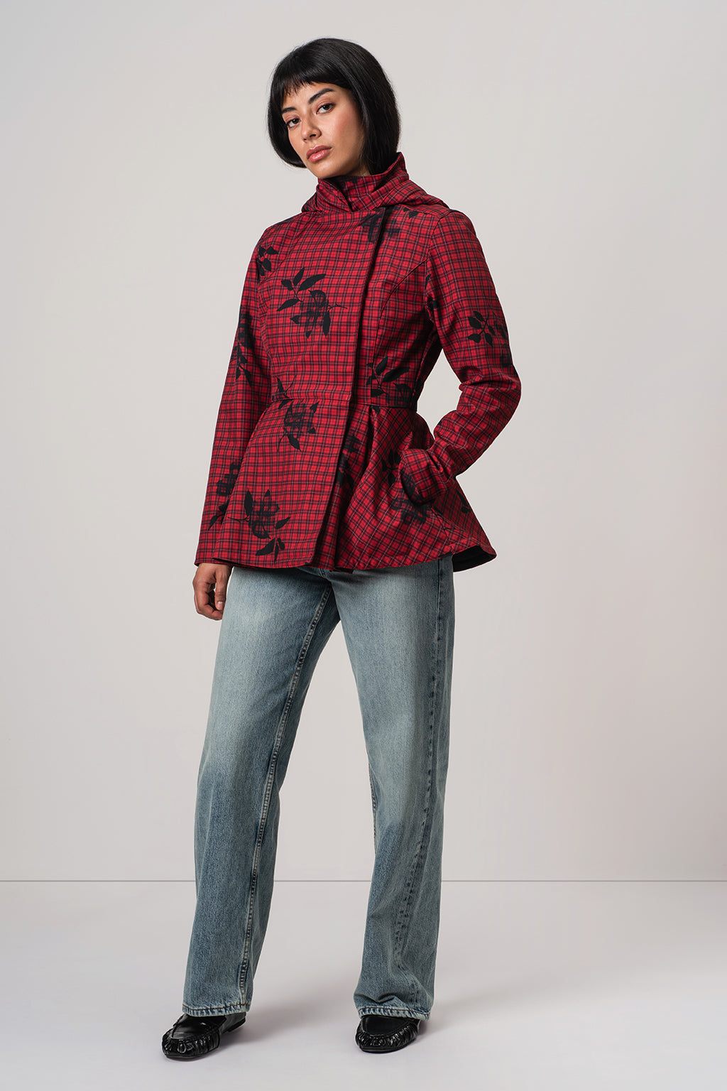 A model wearing RainSisters red and black checkered hooded jacket Crimson Heart with black floral print, paired with light blue jeans and black shoes, standing against a light gray studio background.