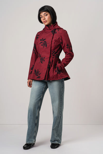 A model wearing RainSisters red and black checkered hooded jacket Crimson Heart with black floral print, paired with light blue jeans and black shoes, standing against a light gray studio background.