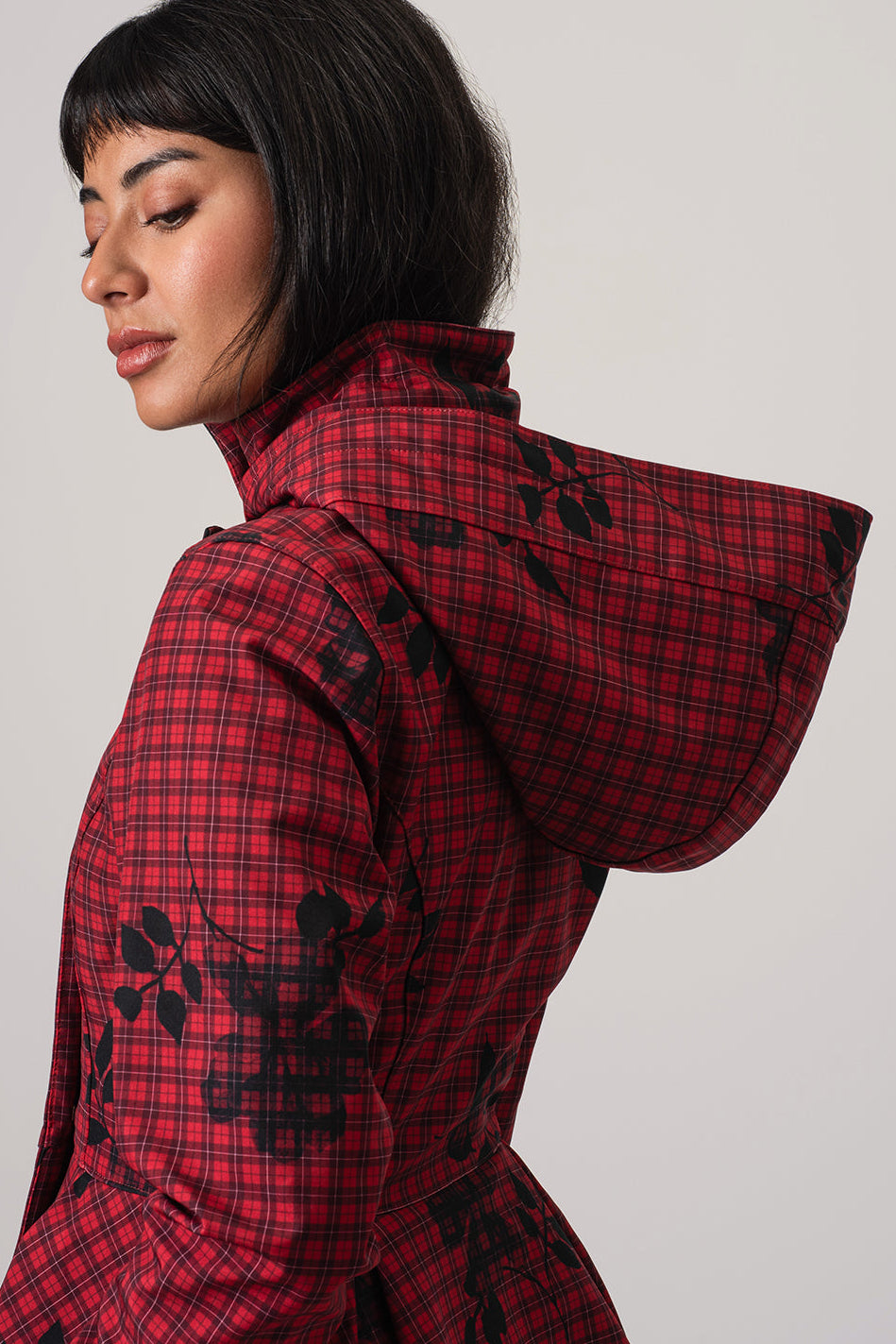 A model shown from the side and back wearing RainSisters red and black plaid hooded jacket Crimson Heart with black floral pattern details and detachable hood, set against a plain light gray studio background.