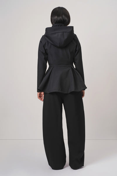 A model viewed from the back wearing RainSisters black hooded jacket Moonlit Bow with a peplum waist and wide-leg black pants, set against a plain light gray studio background.