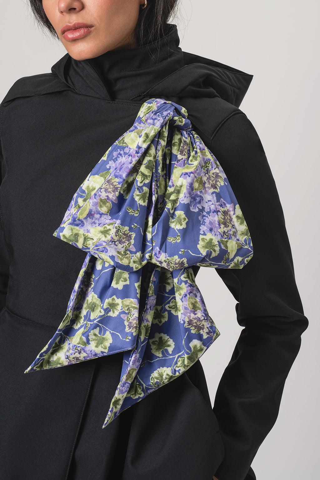 A close-up of a model wearing RainSisters black waterproof jacket Moonlit Bow with detachable large, draped floral scarf in purple, green, and blue tones.