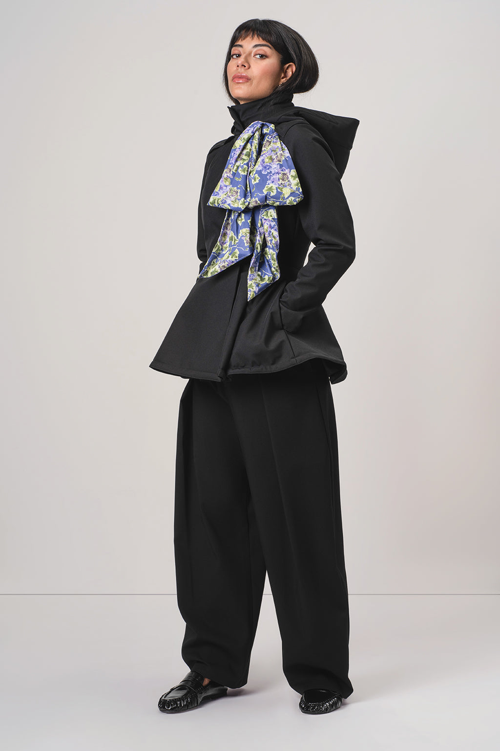 A model wearing RainSisters black hooded jacket Moonlit Bow with detachable large blue and green patterned bow at the collar, paired with black wide-leg trousers and a black beret, standing against a light gray studio background.