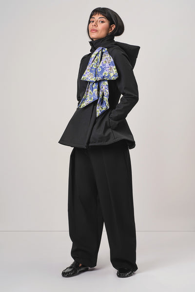 A model wearing RainSisters black hooded jacket Moonlit Bow with detachable large blue and green patterned bow at the collar, paired with black wide-leg trousers and a black beret, standing against a light gray studio background.