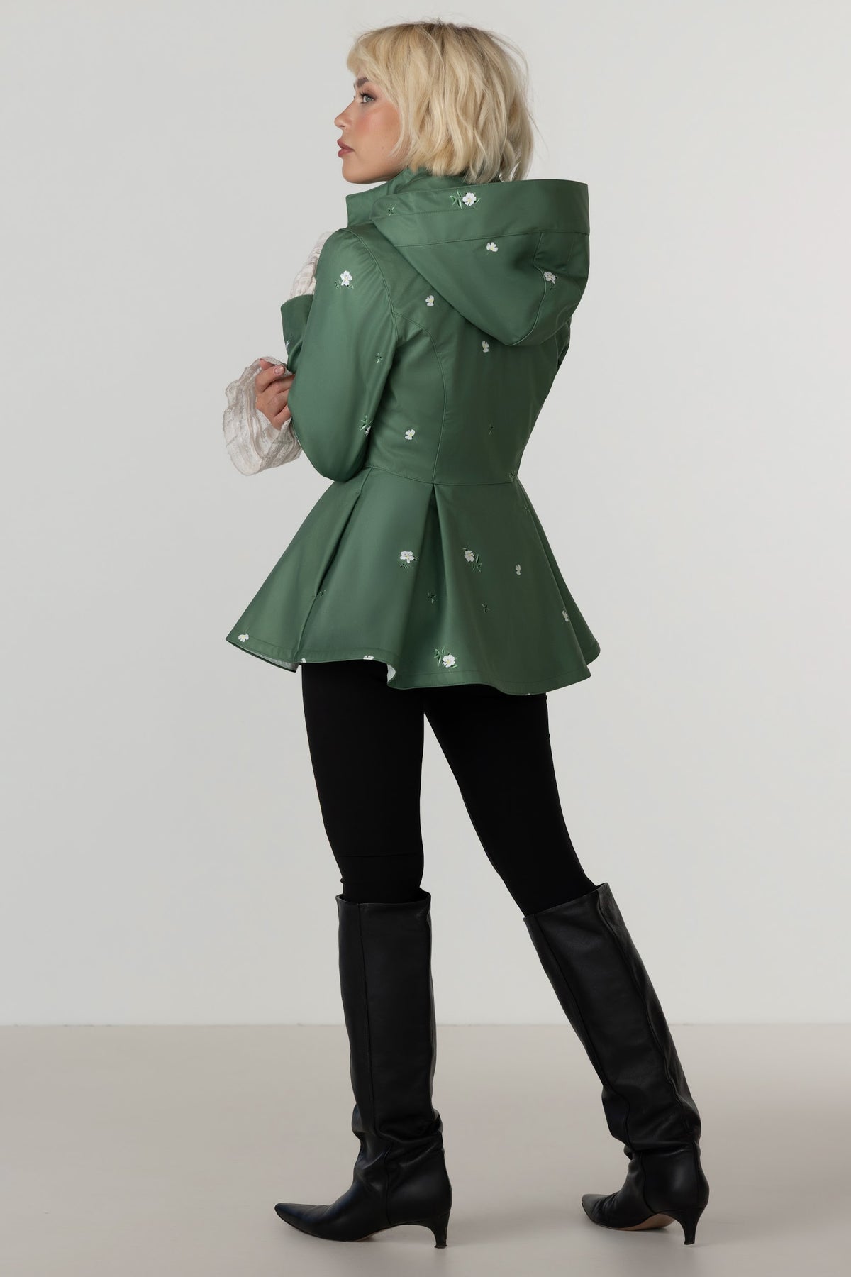 A woman modeling RainSisters green hooded raincoat Primrose Green with white flower details, paired with black leggings and black knee-high boots, shown from a side-back angle against a light gray studio background.