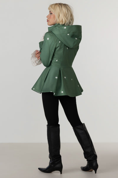 A woman modeling RainSisters green hooded raincoat Primrose Green with white flower details, paired with black leggings and black knee-high boots, shown from a side-back angle against a light gray studio background.