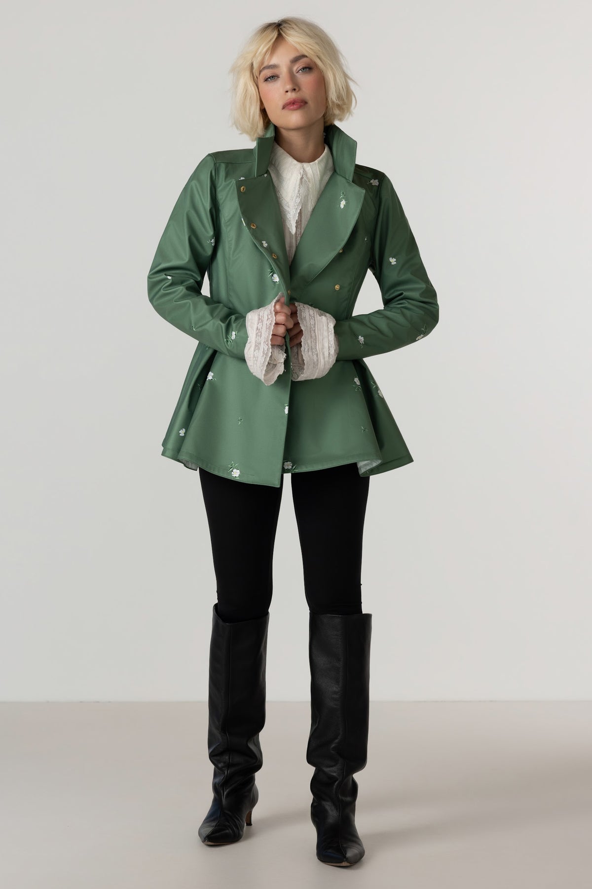 A model stands against a light gray studio background wearing RainSisters green double-breasted jacket Primrose Green with gold buttons, a white ruffled shirt, black pants, and black knee-high boots.