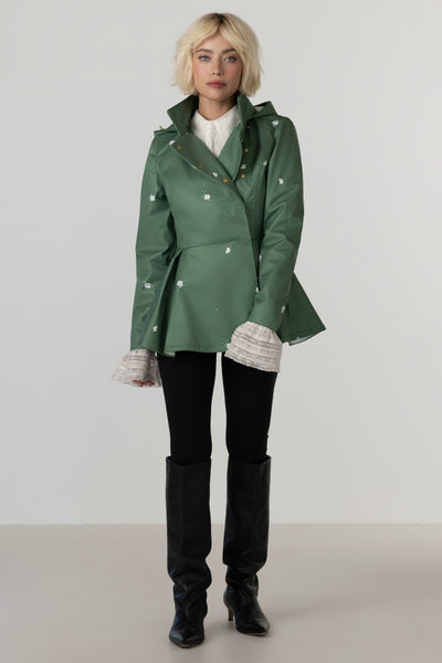 A model wearing RainSisters green double-breasted peplum-style coat Primrose Green with white floral print, styled with a white top, black pants, and black knee-high boots against a neutral studio background.