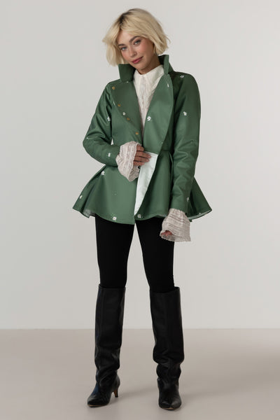 A model wearing RainSisters green  peplum jacket Primrose Green with white flower print, styled over a white ruffled shirt, black pants, and black knee-high boots, set against a plain light gray studio background.