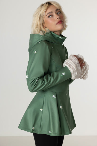 A woman wearing RainSisters sage green hooded raincoat Primrose Green with white flowers, paired with white lace cuffs and black pants, photographed against a light gray studio background.