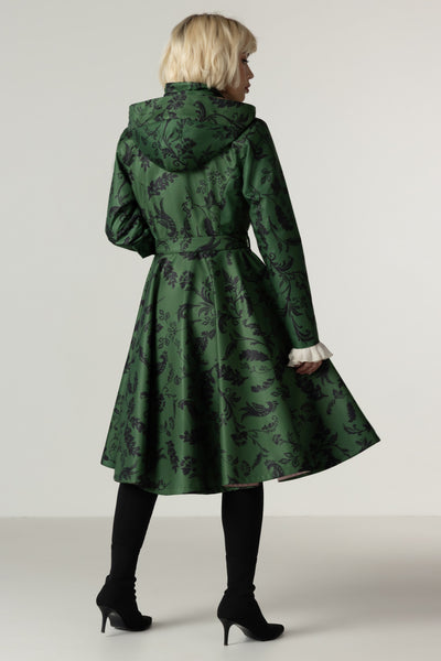 A woman shown from the back wearing a green hooded winter coat with a dark floral print, black tights, and black high heels against a light gray studio background.