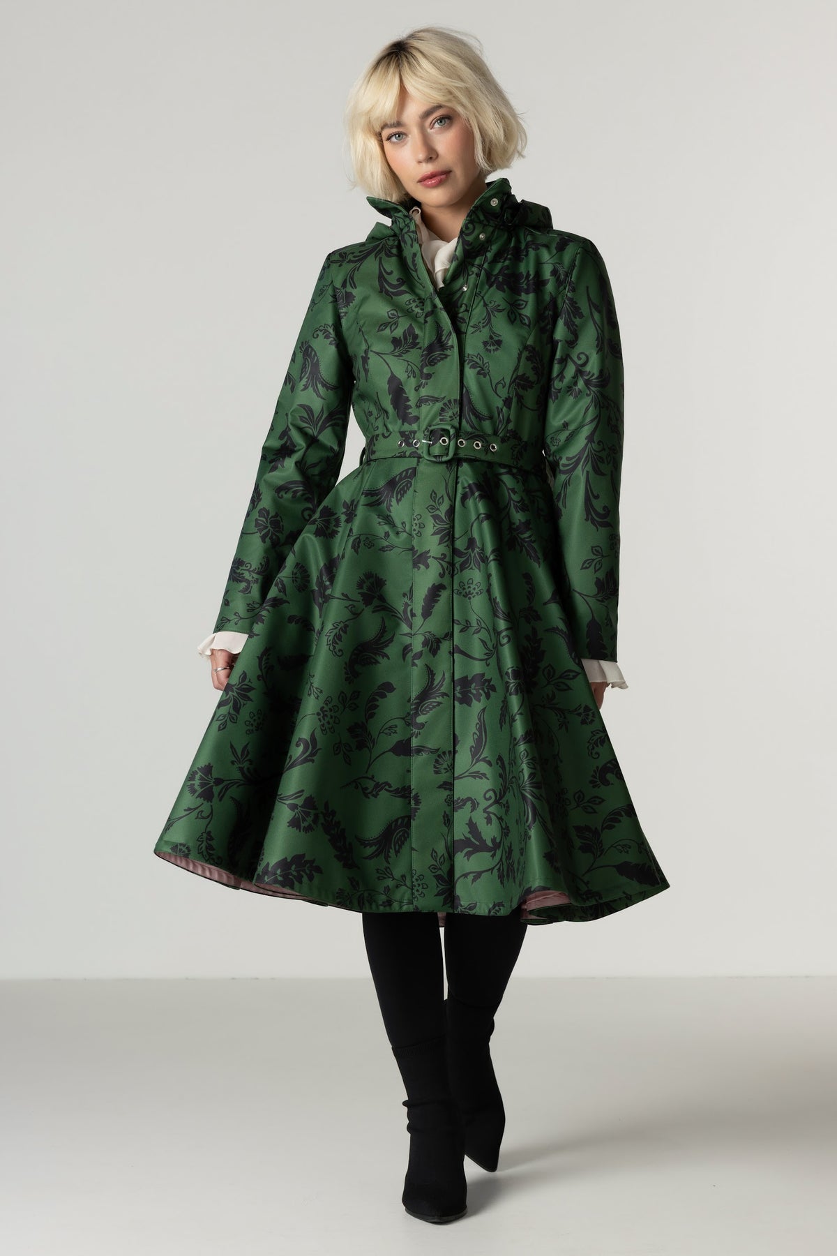 A blonde model wearing a Rainsisters green hooded coat with a black botanical pattern, styled with a belt and black tights against a light gray studio background.