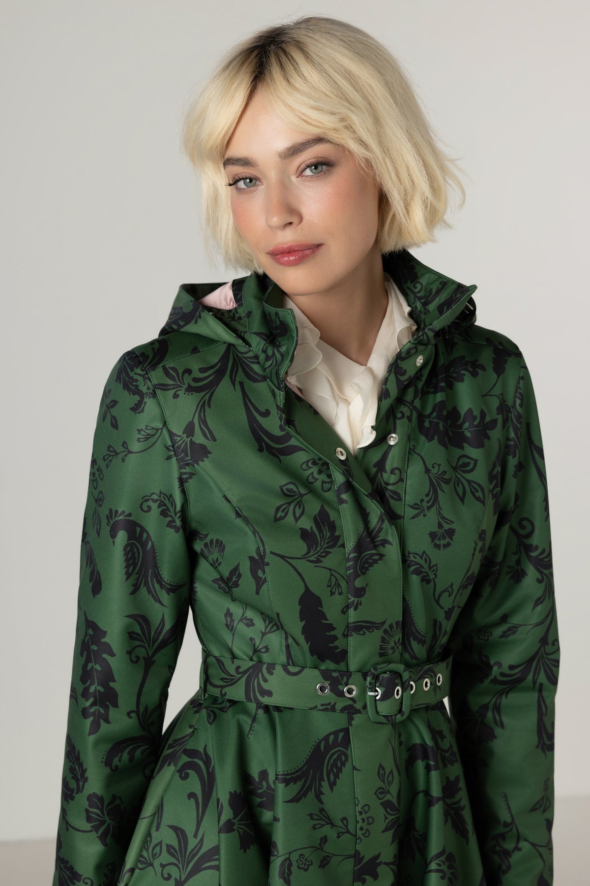 A blonde woman wearing a dark green hooded winter overcoat with a black floral pattern, styled over a cream collared shirt, against a light gray studio background.