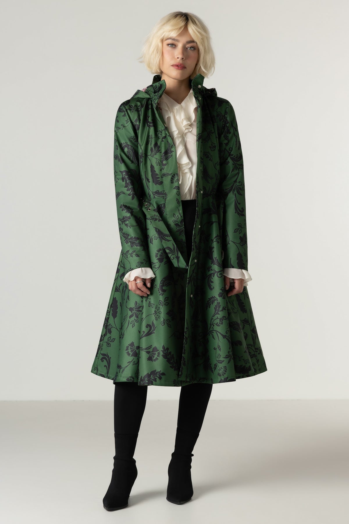 A model wearing a green hooded waterproof winter coat with a dark botanical pattern, styled over a white ruffled blouse, black trousers, and black boots, set against a plain light gray studio background.