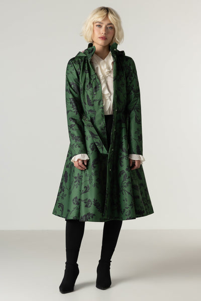 A model wearing a green hooded waterproof winter coat with a dark botanical pattern, styled over a white ruffled blouse, black trousers, and black boots, set against a plain light gray studio background.