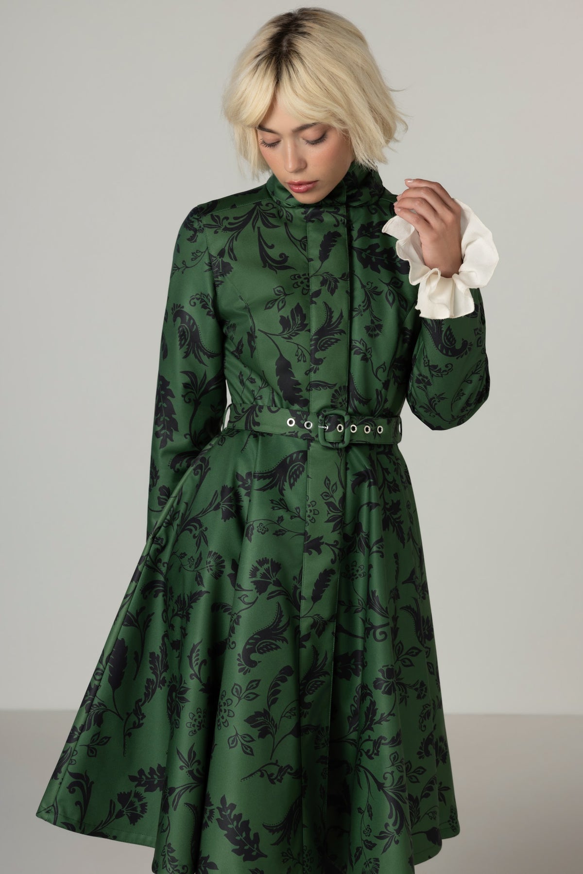 A woman wearing a green long-sleeve midi winter coat with a black floral pattern, a matching belt, and contrasting white ruffled cuffs, photographed against a neutral gray studio background.