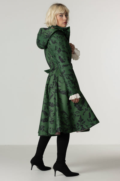 A model wearing a dark green hooded winter coat with a black damask pattern, styled with black tights and black high heels against a plain light gray studio background.