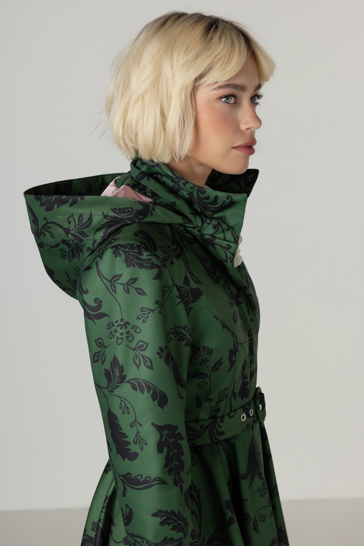 A blonde model shown in profile wearing a dark green hooded Rainsisters winter coat with a black floral pattern, set against a plain light gray studio background.