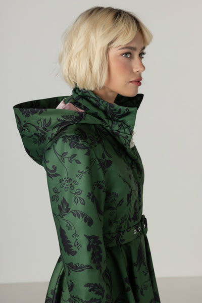 A blonde model shown in profile wearing a dark green hooded Rainsisters winter coat with a black floral pattern, set against a plain light gray studio background.