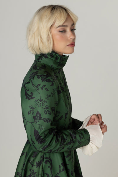 A blonde model in profile wearing a dark green high-neck winter coat with a black floral pattern and contrasting white ruffled cuffs, set against a plain light gray studio background.
