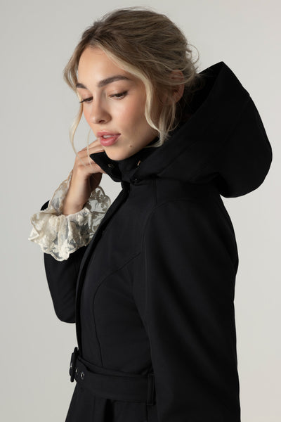 A woman wearing a black Rainsisters hooded winter coat with a belted waist, styled over a white ruffled blouse, photographed against a plain light gray studio background.
