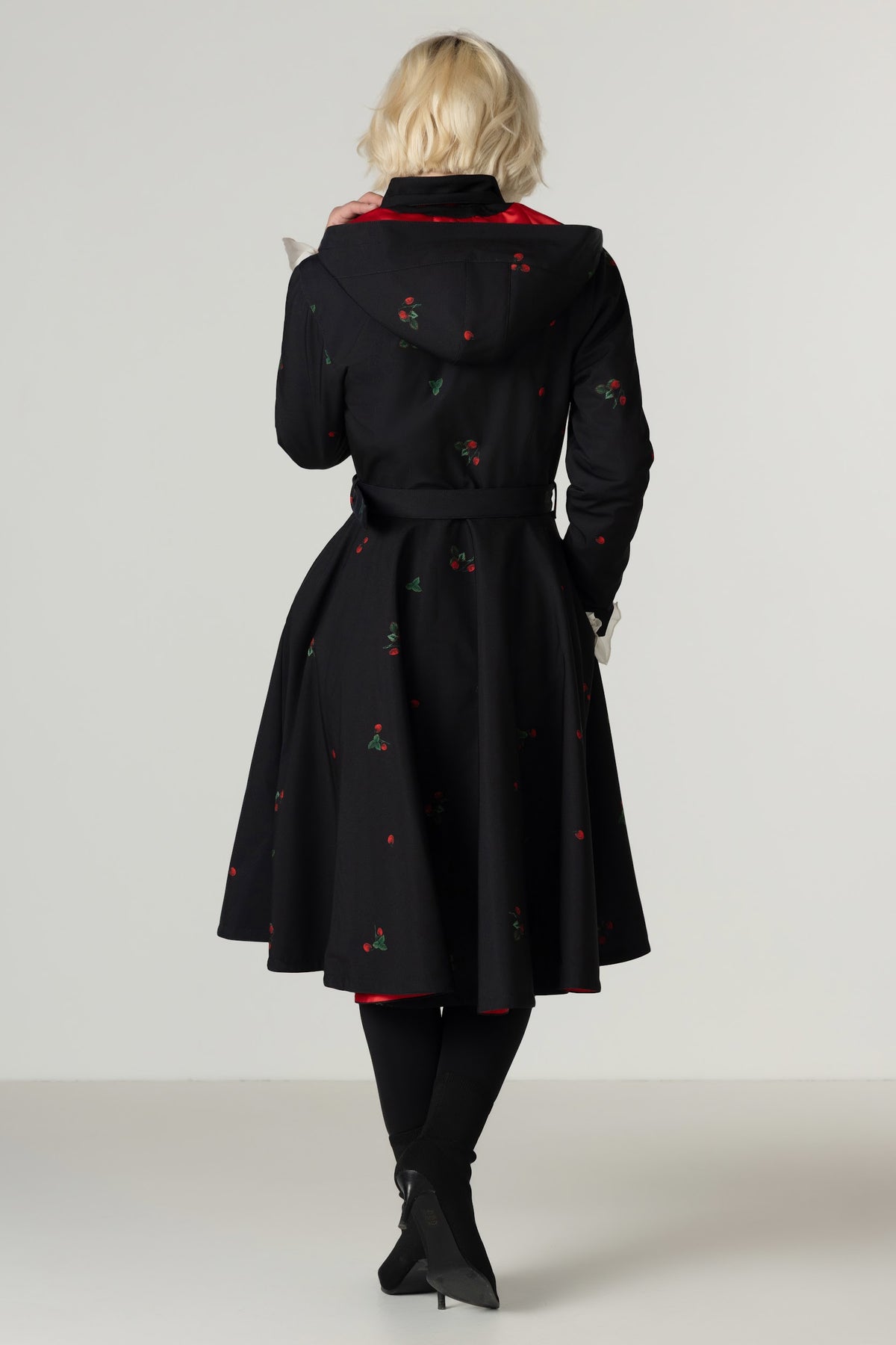 A woman viewed from behind wearing a black hooded winter coat with small red and green berry motifs, paired with black tights and black pointed boots against a light gray studio background.
