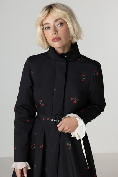 A model wears a black strawberry-print winter coat with a fitted bodice, flared skirt and bold red lining by Rainsisters.