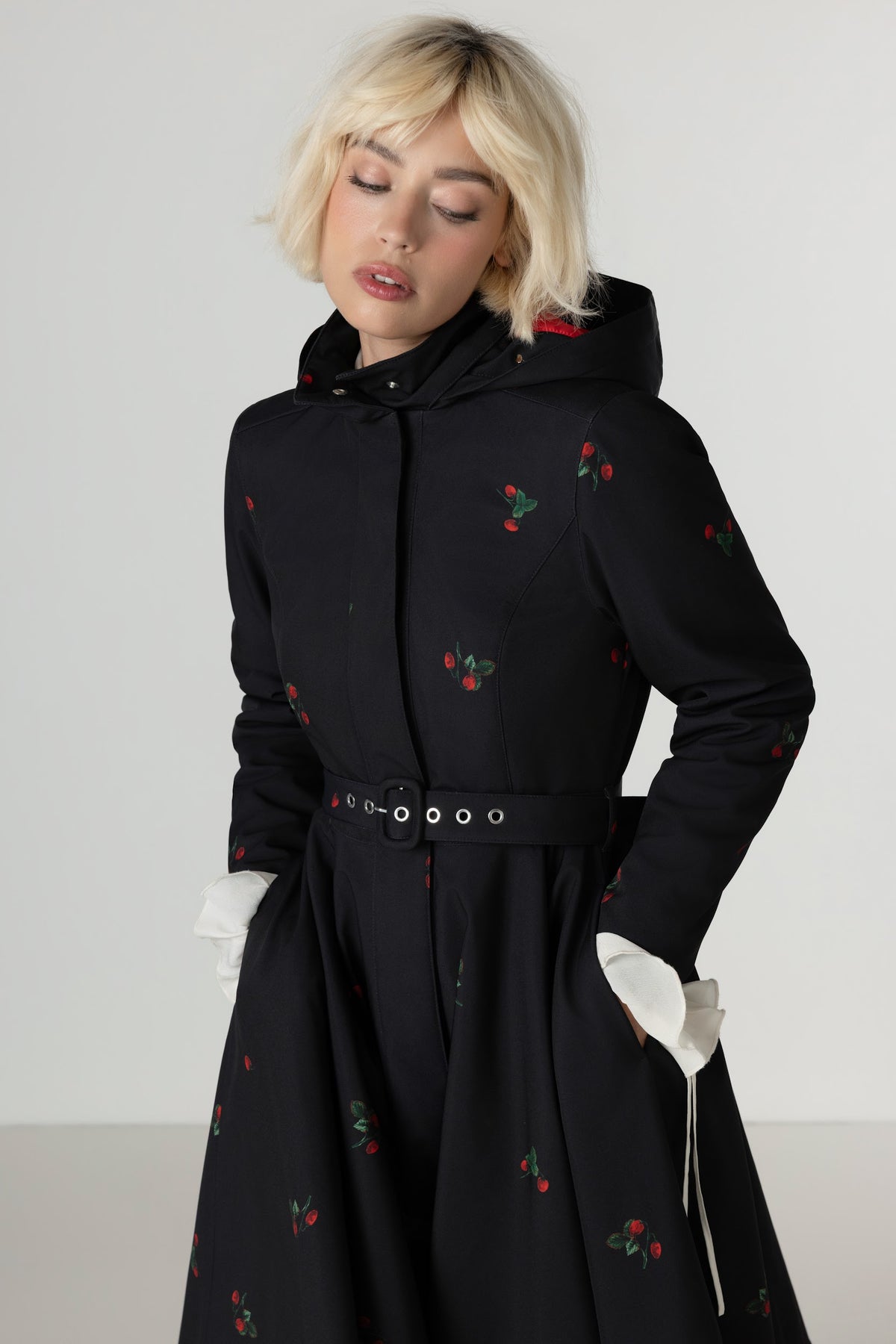 A model with short blonde hair wearing a black hooded winter midi coat with a berry print, and a matching black belt with silver rings, set against a plain light gray background.