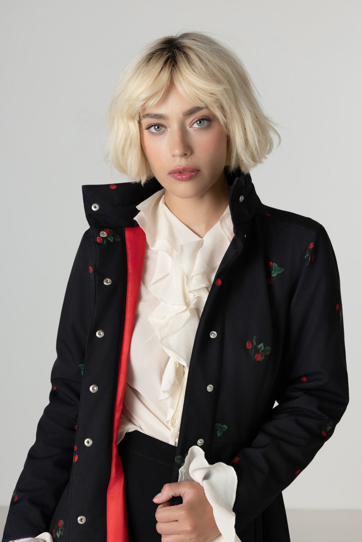 A model with a short blonde bob wearing a black warm winter overcoat with red floral details, layered over a cream ruffled blouse and a bright red inner layer, set against a plain light gray studio background.