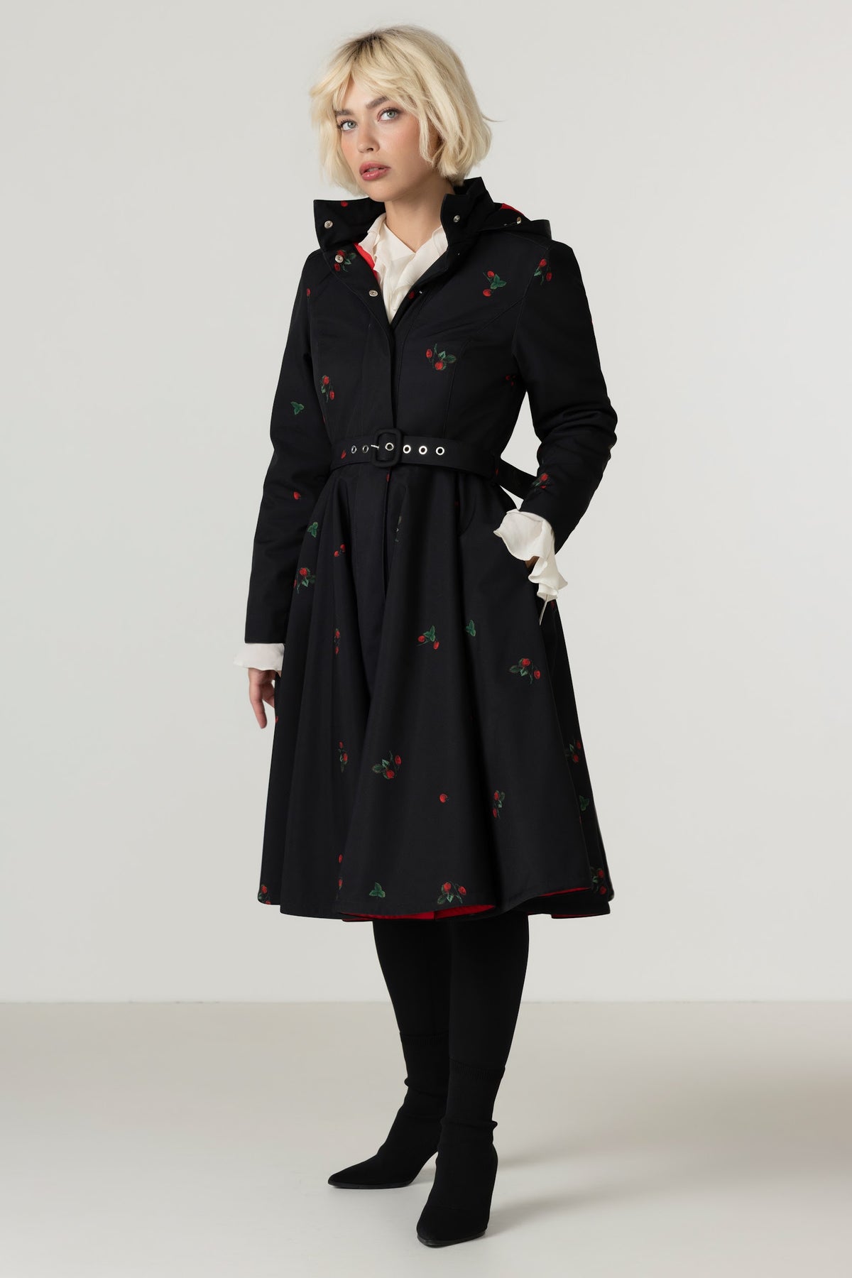 A model wearing a black A-line winter coat with a delicate berry print, a matching belt, and white gloves, standing against a light gray studio background.
