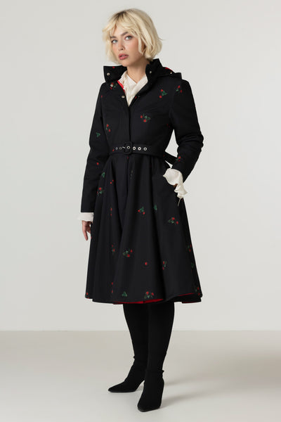 A model wearing a black A-line winter coat with a delicate berry print, a matching belt, and white gloves, standing against a light gray studio background.