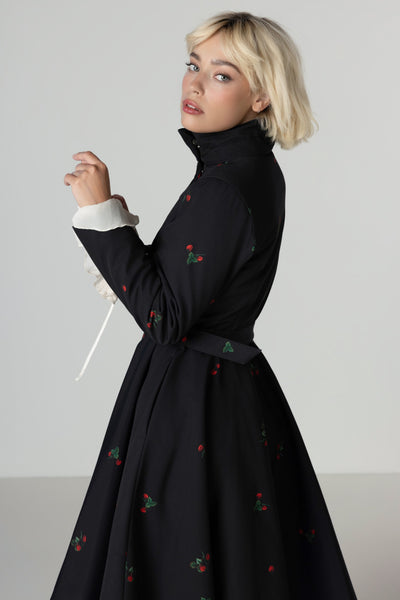 A blonde model wearing a black high-neck long-sleeve winter coat with a matching waist belt and small red floral motifs, photographed against a neutral light gray studio background.