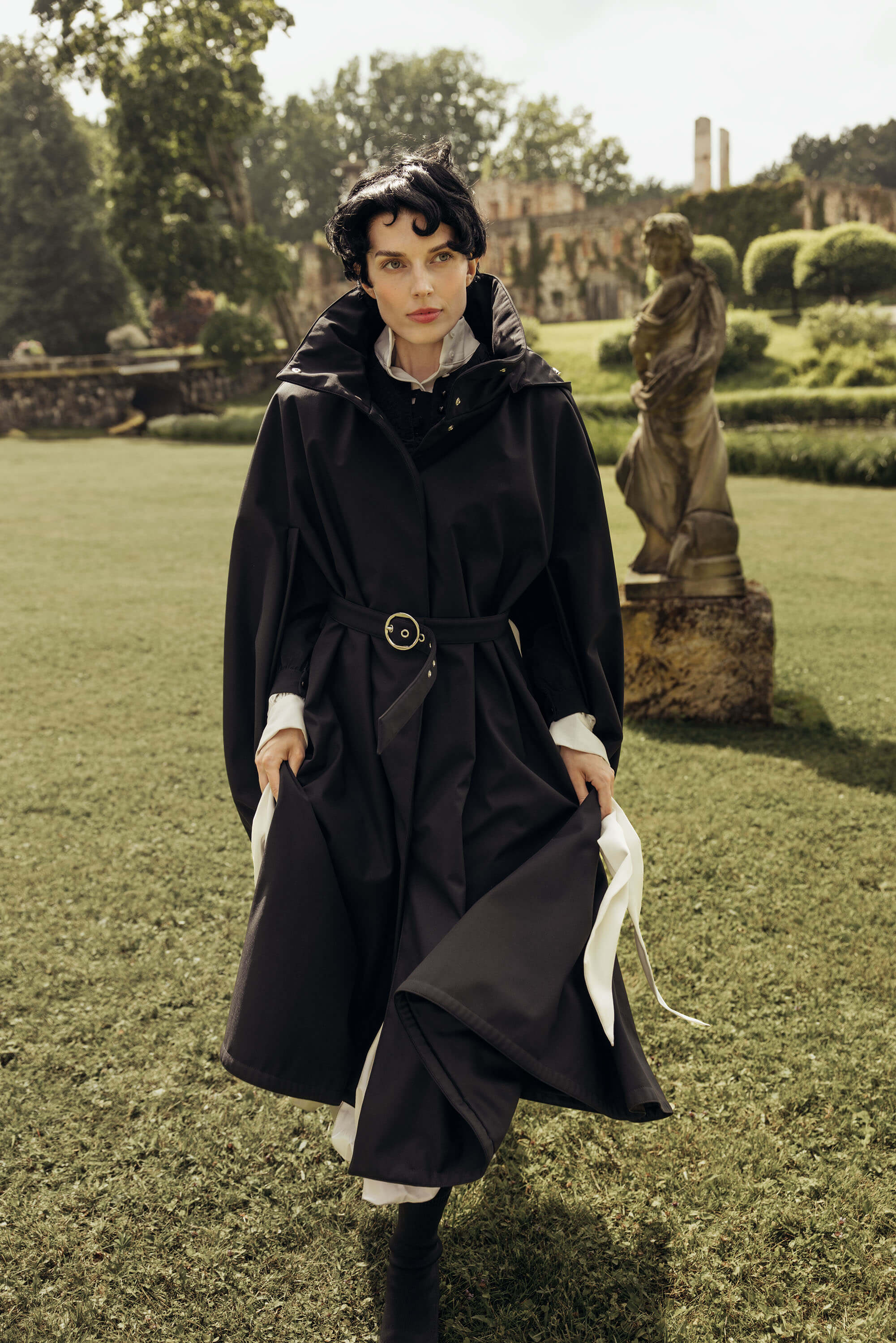 A woman walks on a grassy garden lawn wearing a long black hooded cape coat by RainSisters with a waist belt and styled with white cuffs. A statue and stone building are visible in the background.