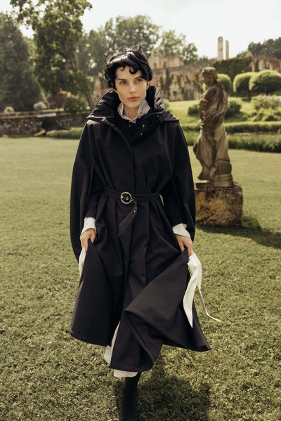 A woman walks on a grassy garden lawn wearing a long black hooded cape coat by RainSisters with a waist belt and styled with white cuffs. A statue and stone building are visible in the background.