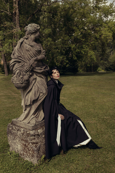 A person in a long black cloak by RainSisters reclines beside a stone statue on a grassy lawn with trees in the background.