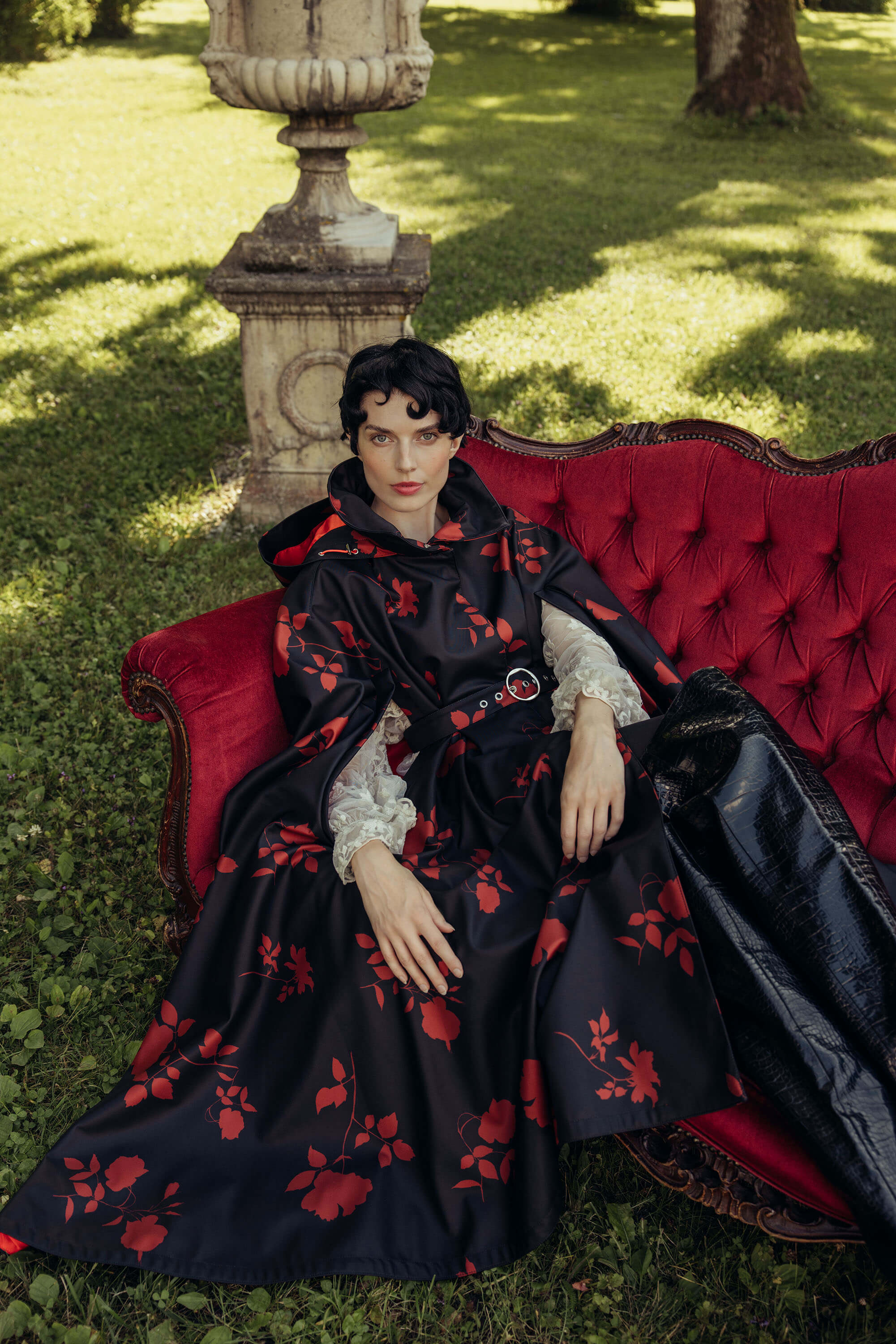 A model wearing a black cloak with red floral pattern by RainSisters styled with white lace sleeves sits on a red tufted outdoor sofa in a grassy garden setting.