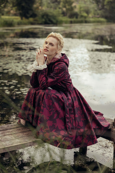 Women's burgundy floral coat with a fitted bodice, full circle skirt, and matching tie belt photographed in a natural outdoor setting.