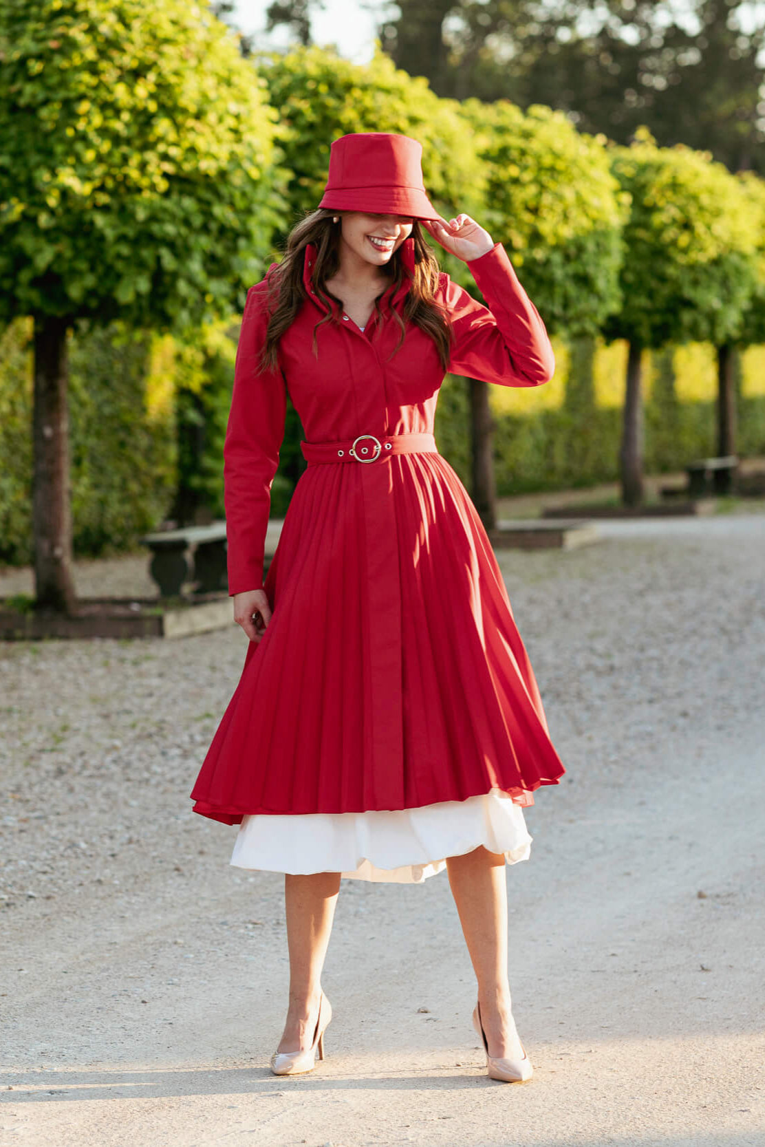 Woman in a red coat and hat standing outdoors with greenery in the background