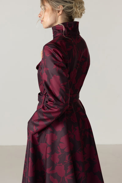 A woman shown from the side and back wearing a dark burgundy and dark  brown patterned raincoat with a high collar and waist tie by RainSisters, photographed against a plain light gray studio background.