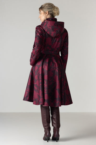 A woman viewed from the back wearing a dark red and black patterned hooded coat with a waist belt by RainSisters, paired with dark brown knee-high boots, against a light gray studio background.