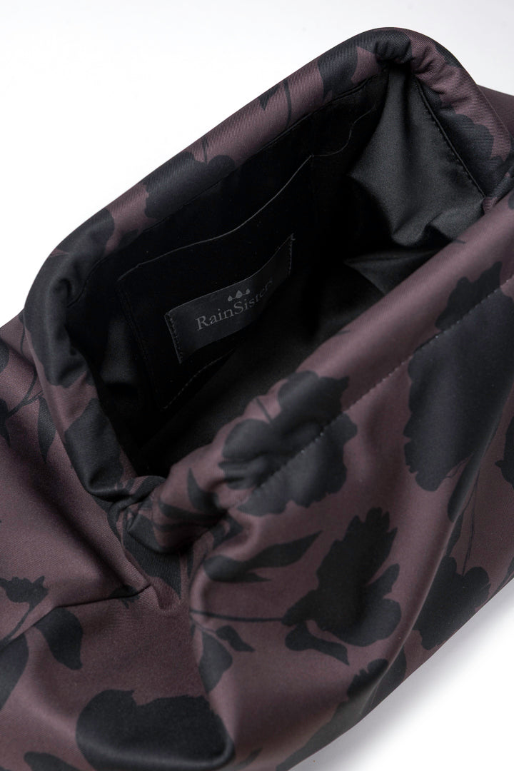 Close-up of a Rainsisters clutch bag with a dark floral pattern in brown with black floral print, showing the black interior lining and a brand label.
