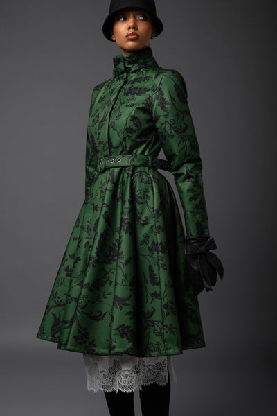 A model wears a dark green high-neck coat with a black floral pattern, styled with a matching belt, black gloves, a black hat, and a white lace hem, set against a dark gray studio background.