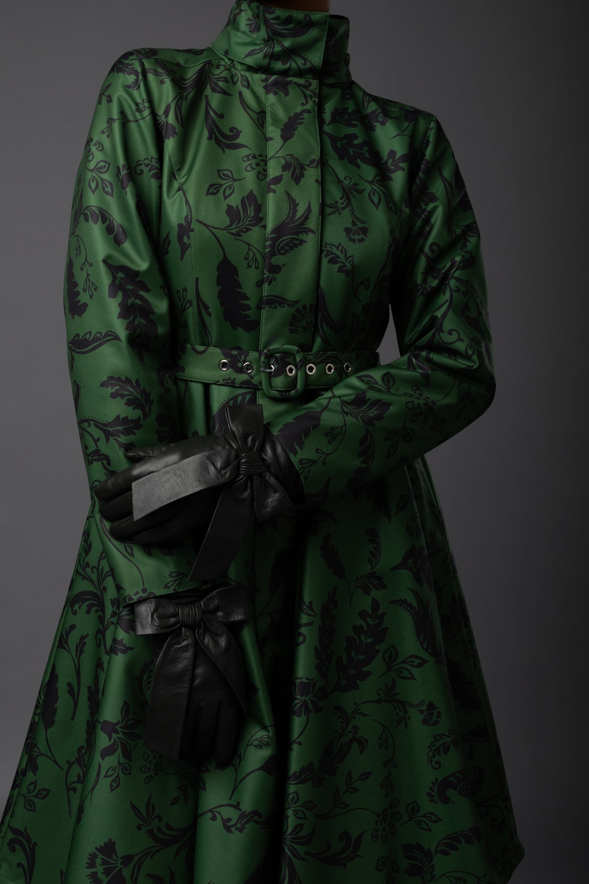 A dark green winter coat with a black floral pattern, featuring a high mock neck, a matching belt with silver-tone studs, and black gloves with ribbon accents, shown against a dark gray studio background.