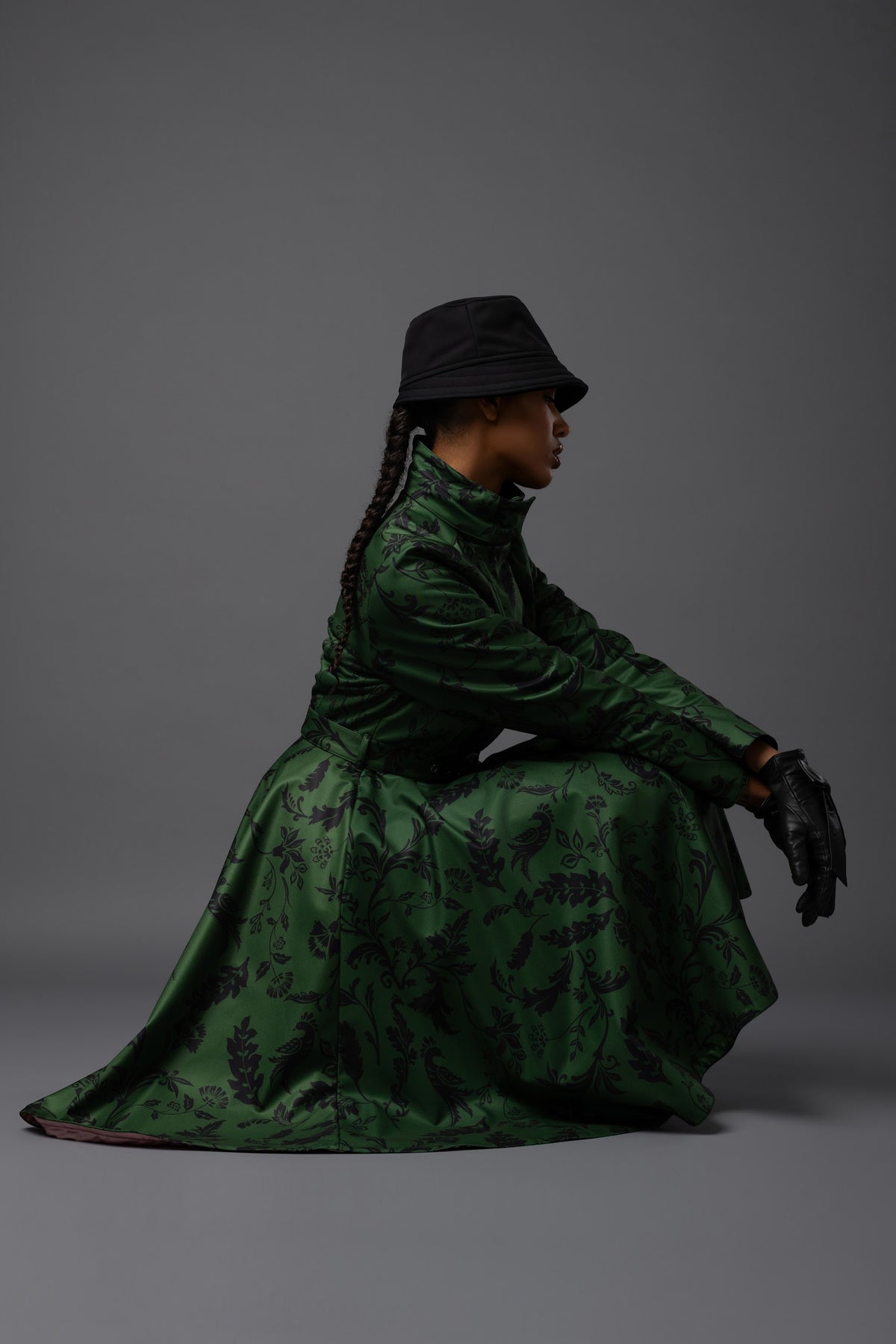 A model wearing a dark green patterned warm winter coat with a black hat, black gloves, posed in profile against a solid dark gray studio background.