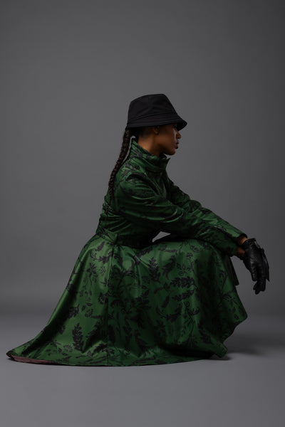 A model wearing a dark green patterned warm winter coat with a black hat, black gloves, posed in profile against a solid dark gray studio background.