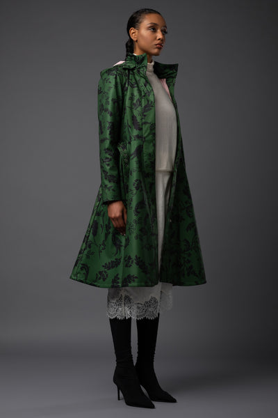 A model is shown from the side wearing Rainsisters long green waterproof coat with a black floral pattern, layered over a light dress, and black knee-high boots against a dark gray studio background.