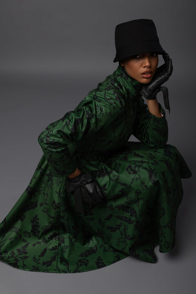 A model wearing a dark green long-sleeved waterproof winter coat with a black floral pattern, a black bucket hat, and black gloves, posed against a dark gray studio background.