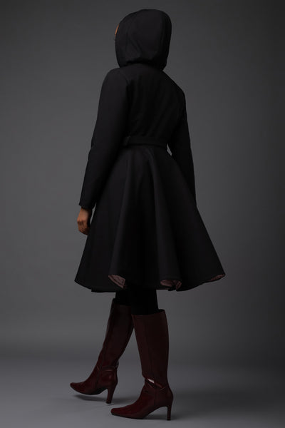 A model from behind wearing a black hooded winter longline Rainsisters coat with a flared skirt and a matching belt against a dark gray studio background.
