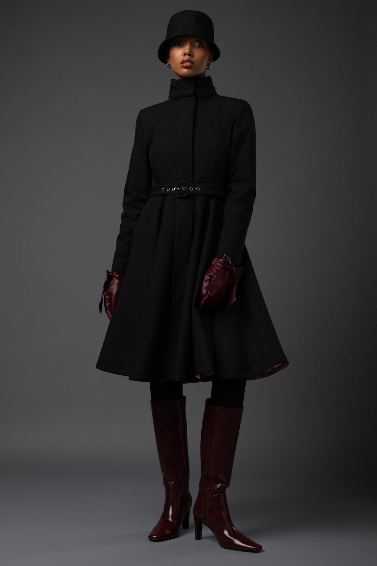 A model wearing a black high-neck long waterproof winter coat with a matching belt, black hat, burgundy gloves, and burgundy knee-high boots, photographed against a dark gray studio background.