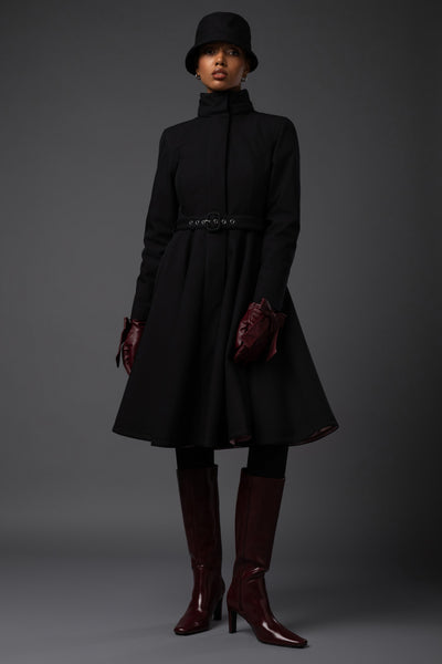 A model wearing a black high-neck long waterproof winter coat with a matching belt, black hat, burgundy gloves, and burgundy knee-high boots, photographed against a dark gray studio background.